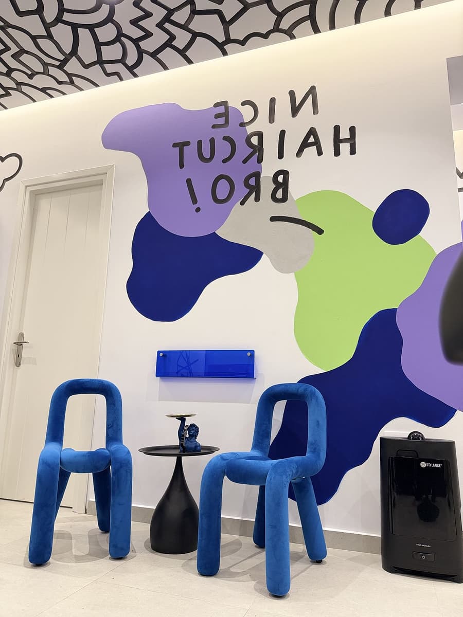 Η φράση NICE HAIRCUT BRO! ζωγραφισμένη ανάποδα στον τοίχο, πάνω από τις μπλε καρέκλες αναμονής